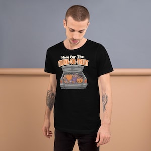 May include: A black short-sleeved t-shirt featuring a graphic of an open car trunk filled with pumpkins and candy. Above the car, the text reads Here For The TRUNK-OR-TREAT in white and orange lettering, centered on the chest of the garment.