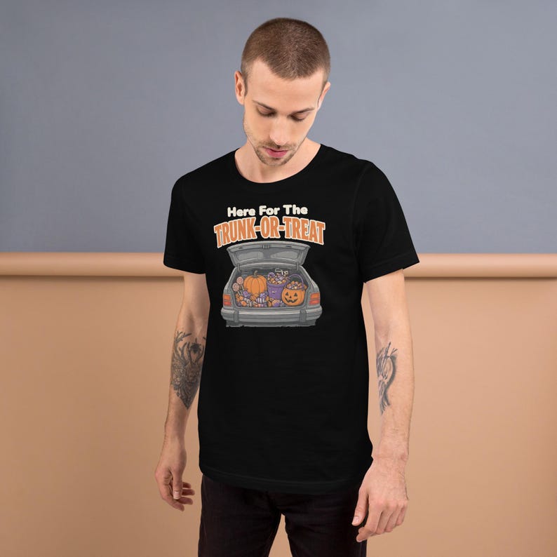 May include: A black short-sleeved t-shirt featuring a graphic of an open car trunk filled with pumpkins and candy. Above the car, the text reads Here For The TRUNK-OR-TREAT in white and orange lettering, centered on the chest of the garment.