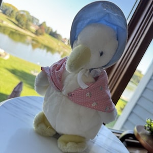 May include: A plush white duck toy wearing a light blue bonnet with a white ribbon tie and a pink cape featuring a small white polka dot and floral pattern. The duck has a soft yellow beak and yellow feet, with the word Jemima embroidered on the bonnet.
