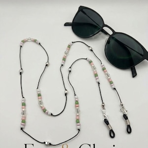 May include: A black cord eyewear chain featuring small pink, green, white, and clear beads interspersed with silver-toned metal accents. The chain includes silver lobster clasps and black rubber loops for attachment to black sunglasses.