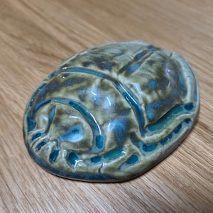 May include: A ceramic scarab beetle figurine featuring a glossy glaze with a blend of earthy olive green and vibrant turquoise blue tones. The surface has a smooth, reflective finish with carved details defining the head, wings, and body.