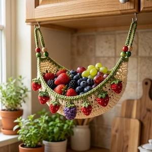 Puede incluir: Una hamaca para frutas de ganchillo color crema con borde verde, colgada de dos ganchos. La hamaca está decorada con pequeñas fresas y una mora de ganchillo colgantes, y llena de fresas frescas, arándanos y uvas verdes.