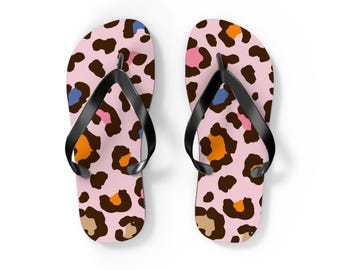 May include: A pair of flip-flops featuring a light pink base with a multi-colored leopard print pattern in shades of dark brown, orange, blue, and tan. Each sandal has a solid black rubber thong strap attached to the footbed.