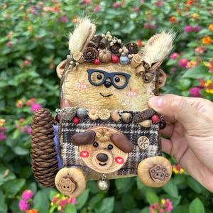May include: A handcrafted animal-shaped ornament featuring a light wood face with black glasses, a brown plaid fabric body, and a pinecone tail. The figure is decorated with small dried flowers, acorns, and berries, and includes a small metal bell at the base.