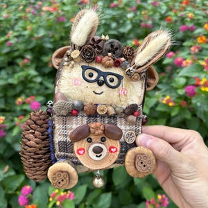 May include: A handcrafted decorative figure featuring a textured, light-colored face with black glasses, adorned with dried pinecones, seeds, and fluffy ears. The body is covered in a brown plaid fabric, accented with a small wooden bear face and a pinecone tail.