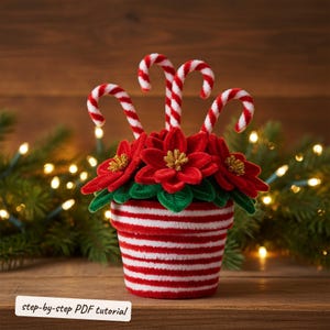 Puede incluir: Una maceta decorativa con rayas horizontales rojas y blancas que contiene flores de pascua rojas con centros dorados y tres tallos de bastones de caramelo a rayas rojas y blancas. El texto step-by-step PDF tutorial es visible abajo.