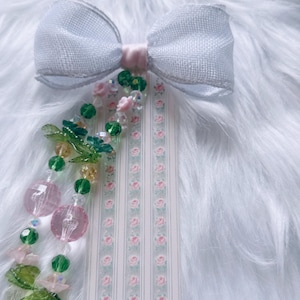 May include: A white fabric bow attached to a rectangular bookmark featuring a vertical striped pattern with small pink roses. Dangling from the centre are two beaded strands with green glass leaves, pink floral beads, and clear round beads.