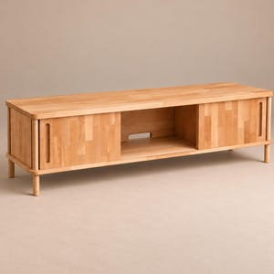 May include: A light-colored wooden media console featuring a rectangular top, four short cylindrical legs, and two sliding cabinet doors with vertical cut-out handles. The center section includes an open shelf with a small rectangular cable cutout.