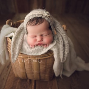 May include: A newborn baby wearing a white knitted hat with bunny ears, sleeping in a woven basket. The baby is wrapped in a white blanket.