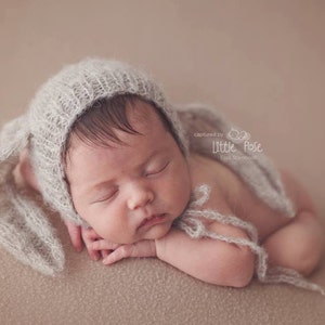 May include: A newborn baby wearing a soft, gray knitted bunny hat with floppy ears. The baby is sleeping peacefully on a light brown background. The hat is made of a delicate, fluffy yarn and has a sweet, whimsical design.