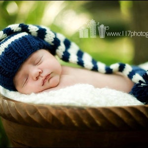 May include: A sleeping baby wearing a blue and white striped knitted hat with a pom-pom on the end. The baby is lying in a wooden bowl.