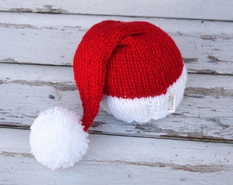 May include: Red and white knitted Santa hat with a white pom-pom. The hat has a white band around the bottom and a white label with the word "Handmade" on it.