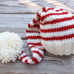 Op de afbeelding: Een rood-wit gestreepte gebreide muts met een lange, puntige top en een witte pompon aan het einde.