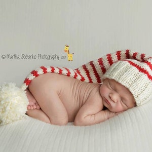 May include: A newborn baby wearing a white and red striped knitted hat with a pom-pom on top. The baby is sleeping on a white blanket.