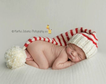 May include: A newborn baby wearing a white and red striped knitted hat with a pom-pom on top. The baby is sleeping on a white blanket.