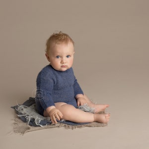 May include: A baby wearing a blue knitted onesie sits on a light brown textured blanket. The baby has blonde hair and blue eyes.