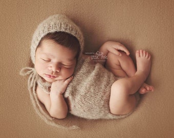 May include: A newborn baby wearing a knitted beige hat and matching outfit is sleeping on a brown background. The baby's eyes are closed and the baby's hand is resting on their cheek. The image is captioned "Captured by Little Pose - Nova Bamboul".