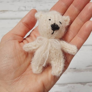 May include: A small, white knitted teddy bear with black eyes and a black nose. The bear is sitting in the palm of a hand.