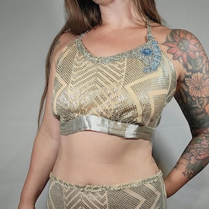 Puede incluir: Un conjunto de dos piezas hecho de tejido de malla dorada con un patrón geométrico. La parte superior es de estilo halter con un ribete de cinta plateada. Los pantalones cortos tienen un ribete blanco.