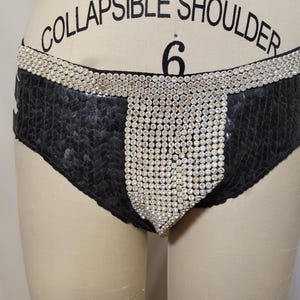 May include: Black sequined bottoms with a wide waistband and a central panel embellished with sparkling rhinestones. The garment is displayed on a mannequin, showcasing its design and texture. The overall aesthetic is glamorous and eye-catching.