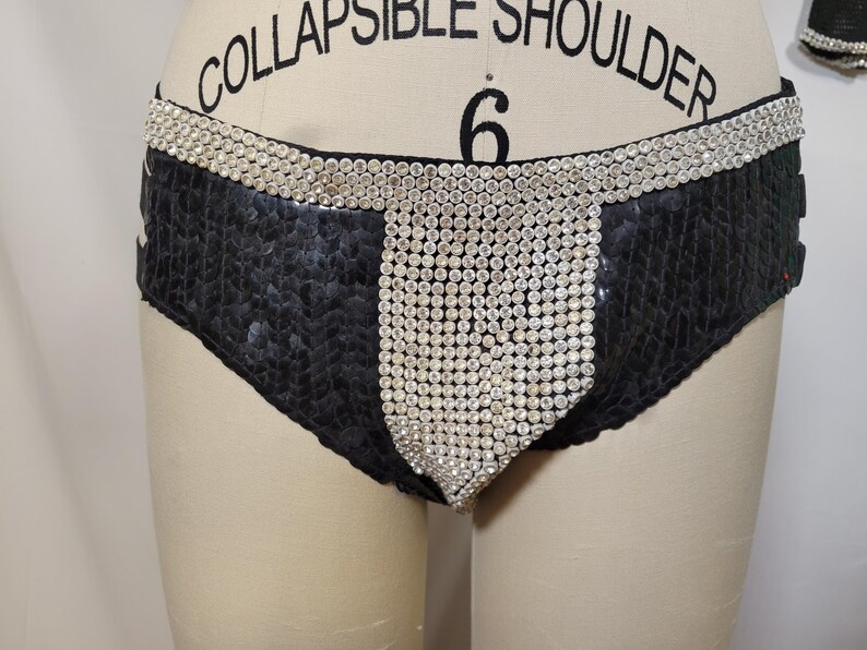 May include: Black sequined bottoms with a wide waistband and a central panel embellished with sparkling rhinestones. The garment is displayed on a mannequin, showcasing its design and texture. The overall aesthetic is glamorous and eye-catching.