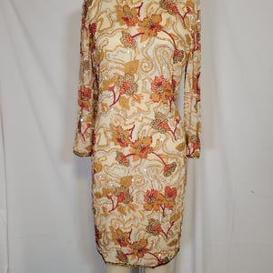 Puede incluir: Vestido color crema de manga larga adornado con lentejuelas doradas, rojas y plateadas en un estampado floral. El vestido presenta un cuello redondo y un dobladillo a la altura de la rodilla. El vestido se exhibe en un maniquí.