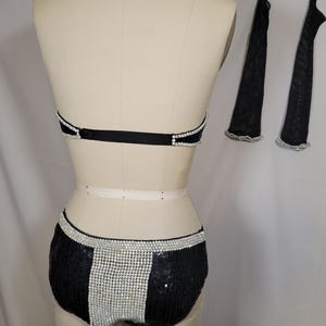 May include: A black sequined dance costume set with silver rhinestone accents. The set includes a halter top, shorts, and two arm sleeves. The shorts have a vertical rhinestone stripe down the center.