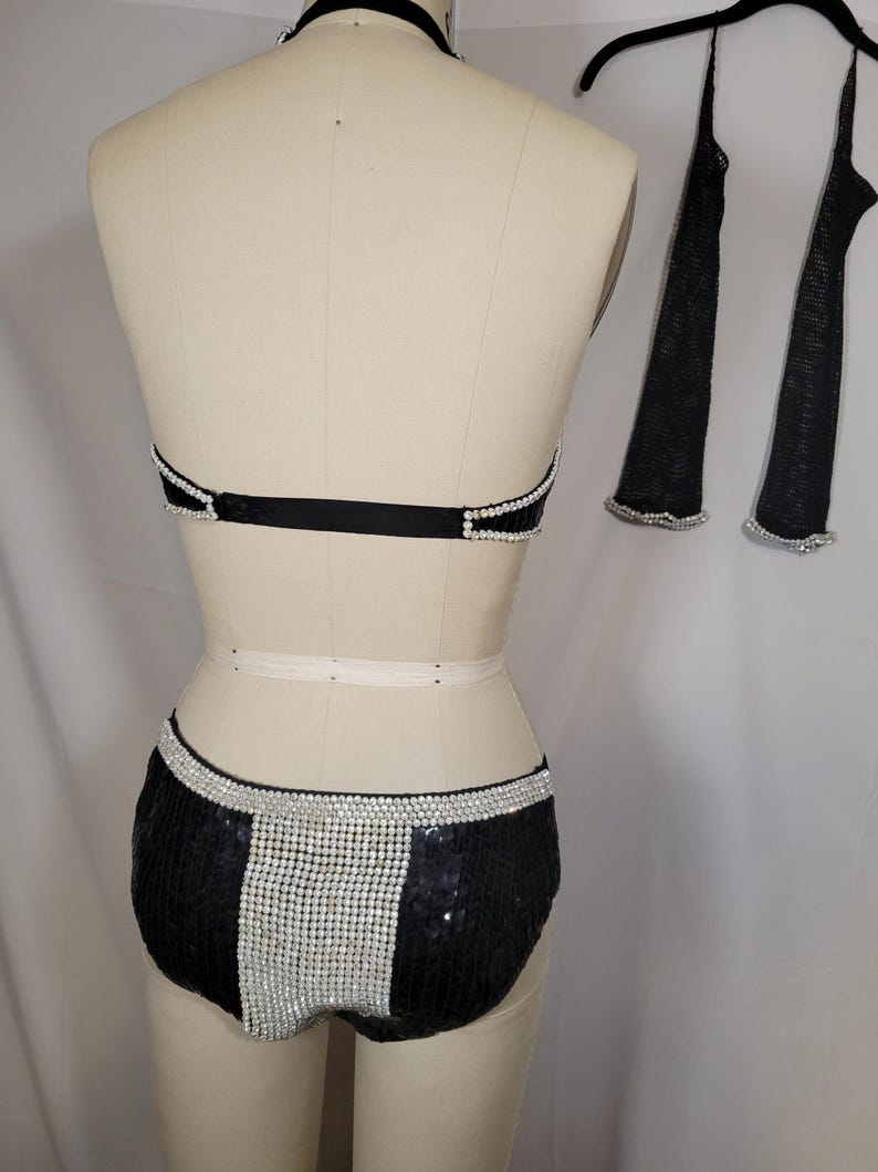 May include: A black sequined dance costume set with silver rhinestone accents. The set includes a halter top, shorts, and two arm sleeves. The shorts have a vertical rhinestone stripe down the center.