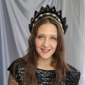 以下が含まれることがあります： 黒いスパンコールのトップスを着た女性が、黒と金のビーズでできたクラウンをかぶっています。クラウンは尖った形をしていて、パールとラインストーンで飾られています。