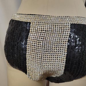 May include: Black sequined shorts with a wide waistband and a vertical panel of sparkling rhinestones. The shorts are designed with a high-waisted cut and are likely part of a costume or performance outfit.