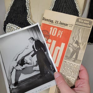 May include: Black halter top with pearl embellishments, vintage photographs, and a newspaper clipping. The photographs feature acrobatic performers. The newspaper is in German and dated January 22.