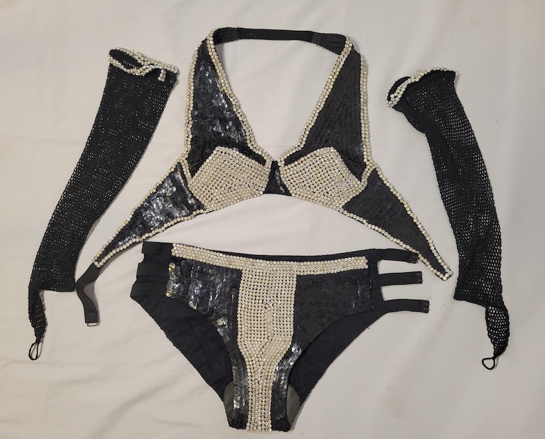 May include: A black sequined bikini set with pearl accents, including a halter top, high-waisted bottoms, and two long gloves. The bikini top and bottoms feature pearl detailing. The gloves are made of a black mesh material.