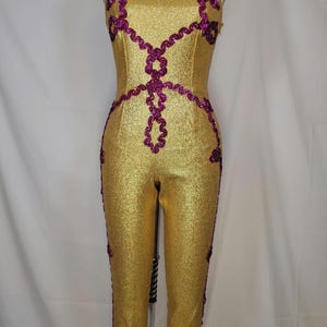 May include: Gold jumpsuit with a purple sequined design. The jumpsuit has a square neckline and a sleeveless design.