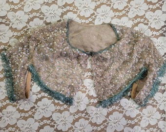May include: A vintage, open-front bolero jacket. The jacket is a light tan color, covered in sequins and beads. The edges are trimmed with a teal fringe. The neckline is a rounded square shape.