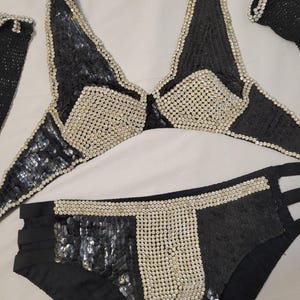 May include: Black sequined bikini set with white beaded accents. The halter top and bikini bottoms feature a geometric design with a mix of sequins and beads. The set includes a matching accessory.