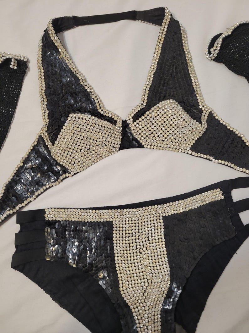 May include: Black sequined bikini set with white beaded accents. The halter top and bikini bottoms feature a geometric design with a mix of sequins and beads. The set includes a matching accessory.