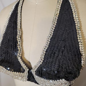 May include: Black sequined halter top with a silver rhinestone trim. The top is displayed on a mannequin. The halter top is a triangle shape and has a clasp closure. The sequins are small and closely spaced.