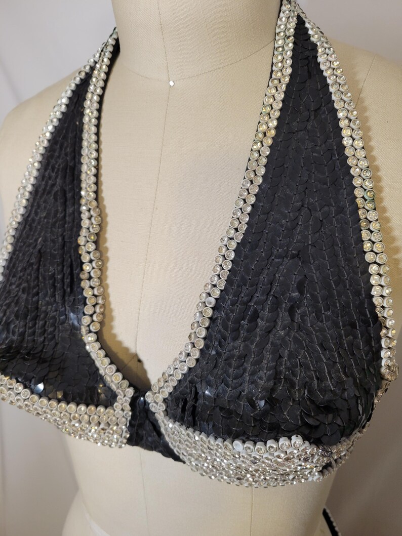 May include: Black sequined halter top with a silver rhinestone trim. The top is displayed on a mannequin. The halter top is a triangle shape and has a clasp closure. The sequins are small and closely spaced.
