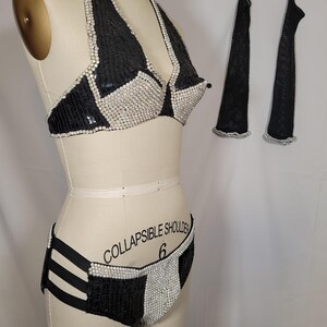 May include: A black and silver sequined bikini set with matching arm covers. The bikini top is a halter style, and the bottoms have a high-waisted design. The arm covers are long and sheer. The set is displayed on a mannequin.
