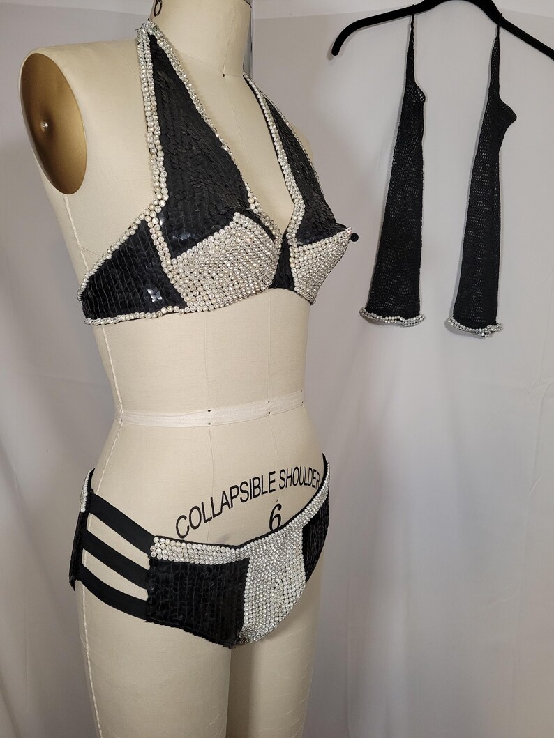 May include: A black and silver sequined bikini set with matching arm covers. The bikini top is a halter style, and the bottoms have a high-waisted design. The arm covers are long and sheer. The set is displayed on a mannequin.
