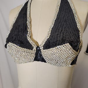 May include: Black halter top with a textured fabric and a sparkling rhinestone trim. The bra-style top is displayed on a mannequin. The top is a dark color with a shiny, textured appearance.