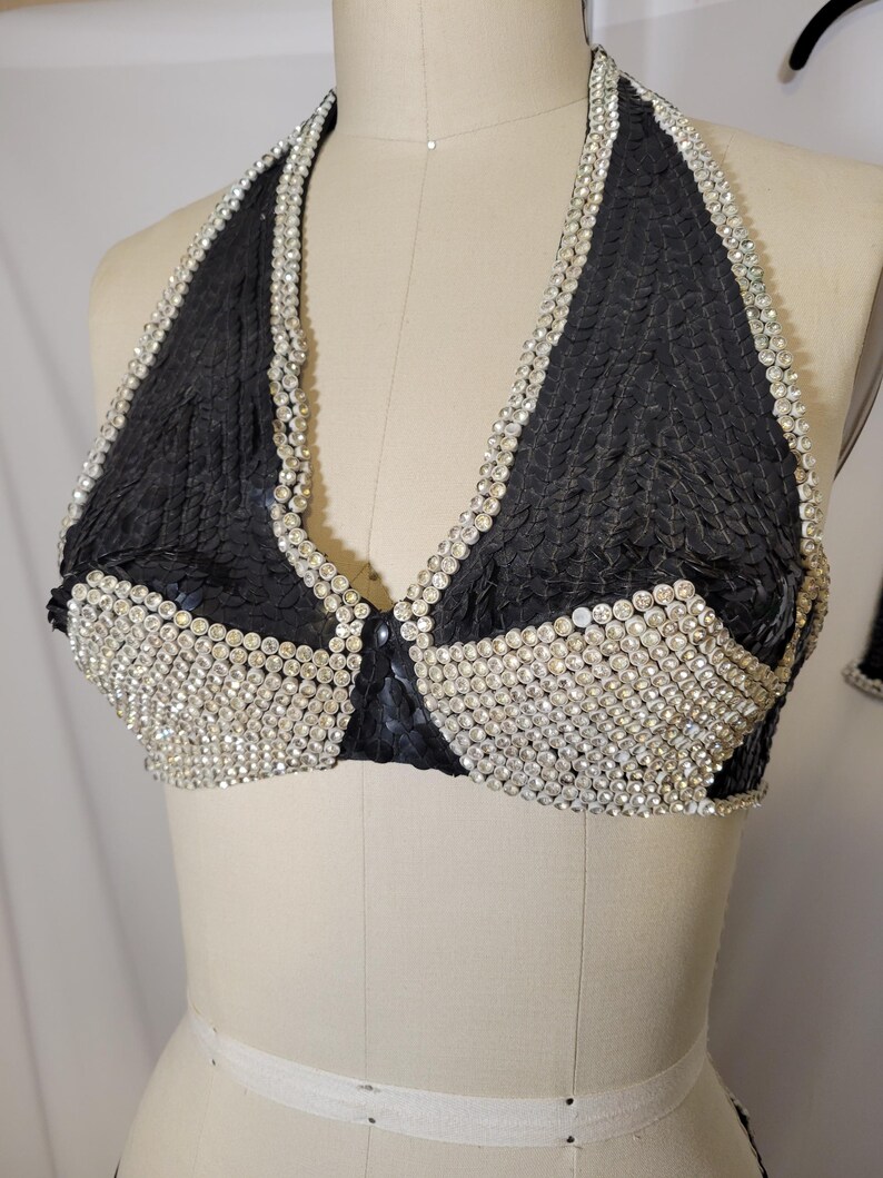 May include: Black halter top with a textured fabric and a sparkling rhinestone trim. The bra-style top is displayed on a mannequin. The top is a dark color with a shiny, textured appearance.