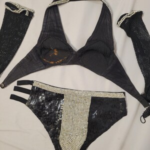 May include: A black lingerie set with a halter top, high-waisted bottoms, and two arm sleeves. The halter top and bottoms are embellished with sequins and faux pearls. The arm sleeves are made of a black mesh material and are also embellished with faux pearls.