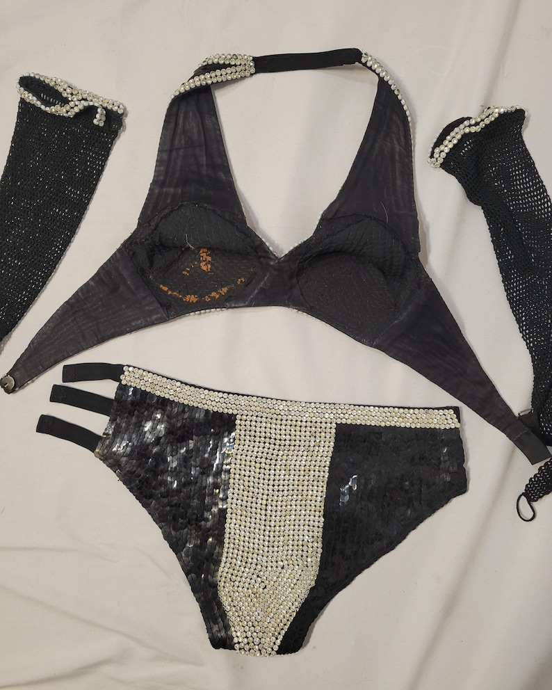 May include: A black lingerie set with a halter top, high-waisted bottoms, and two arm sleeves. The halter top and bottoms are embellished with sequins and faux pearls. The arm sleeves are made of a black mesh material and are also embellished with faux pearls.