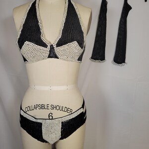 May include: Black halter top bikini set with white embellishments. The bikini top and bottoms are decorated with white beads. Matching black arm covers with white trim are hanging on a black hanger.