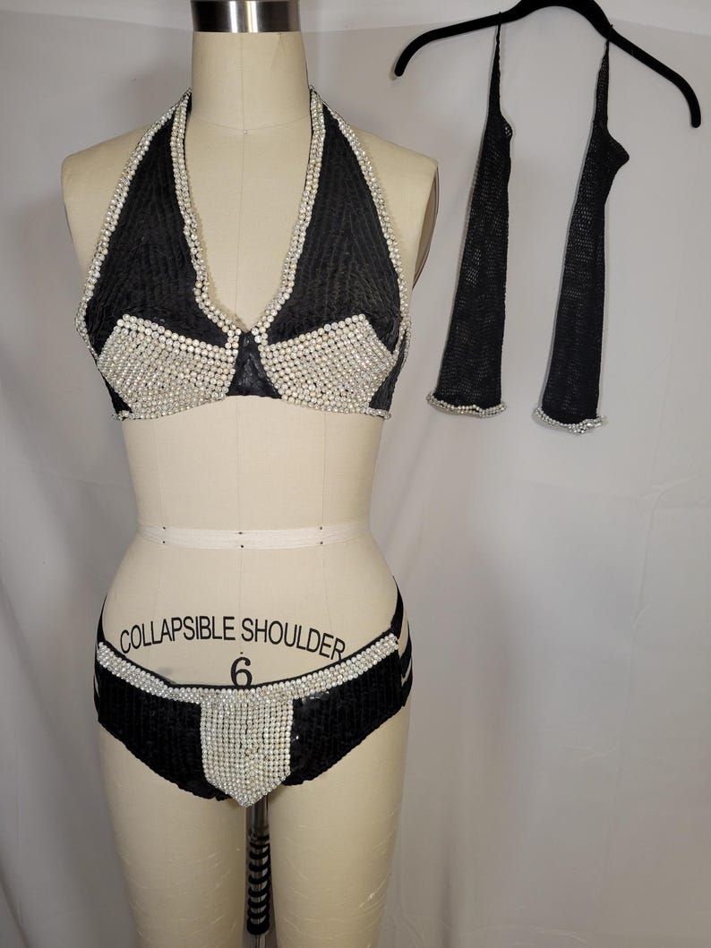 May include: Black halter top bikini set with white embellishments. The bikini top and bottoms are decorated with white beads. Matching black arm covers with white trim are hanging on a black hanger.