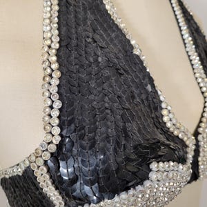 May include: A black sequined halter top with rhinestone trim. The bra-style top features a triangle shape and is embellished with rows of sparkling rhinestones along the edges and straps. The sequins create a textured, shimmering effect.