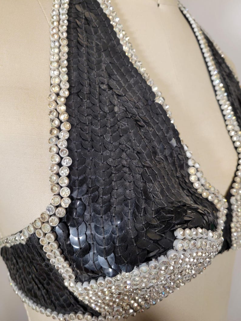 May include: A black sequined halter top with rhinestone trim. The bra-style top features a triangle shape and is embellished with rows of sparkling rhinestones along the edges and straps. The sequins create a textured, shimmering effect.