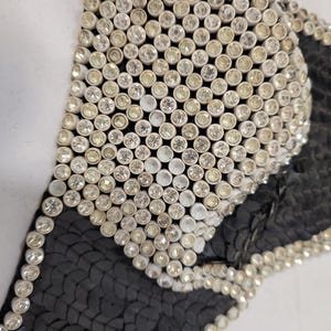 May include: Close-up of a black and silver sequined garment. The garment is heavily embellished with small, round, silver rhinestones. The black fabric is covered in small, black sequins. The design is geometric and appears to be part of a larger piece.