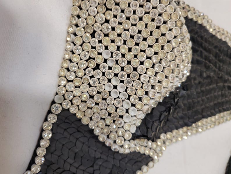 May include: Close-up of a black and silver sequined garment. The garment is heavily embellished with small, round, silver rhinestones. The black fabric is covered in small, black sequins. The design is geometric and appears to be part of a larger piece.